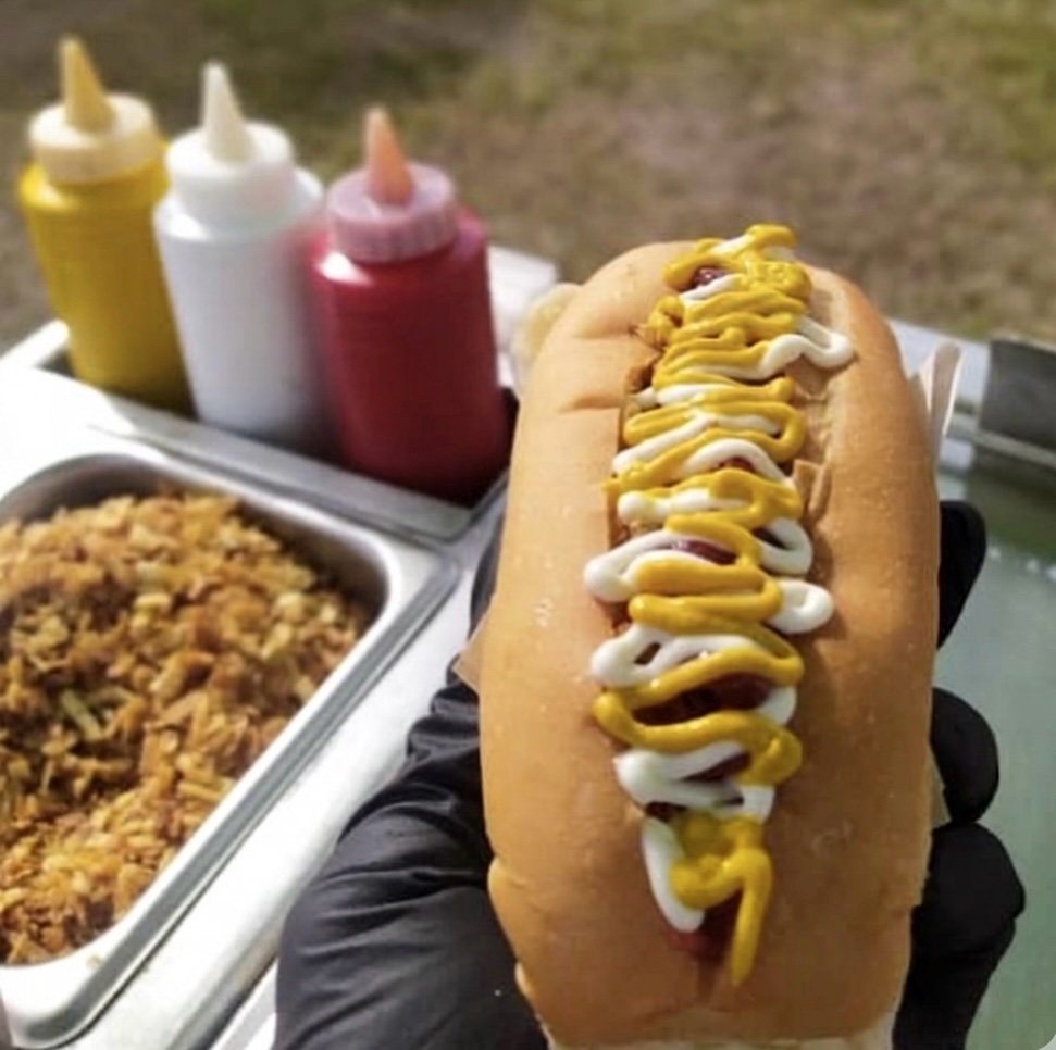 Un perro caliente clásico de Filippo — salchicha al vapor con mostaza, mayonesa y kétchup, con las botellas de la receta original al fondo.